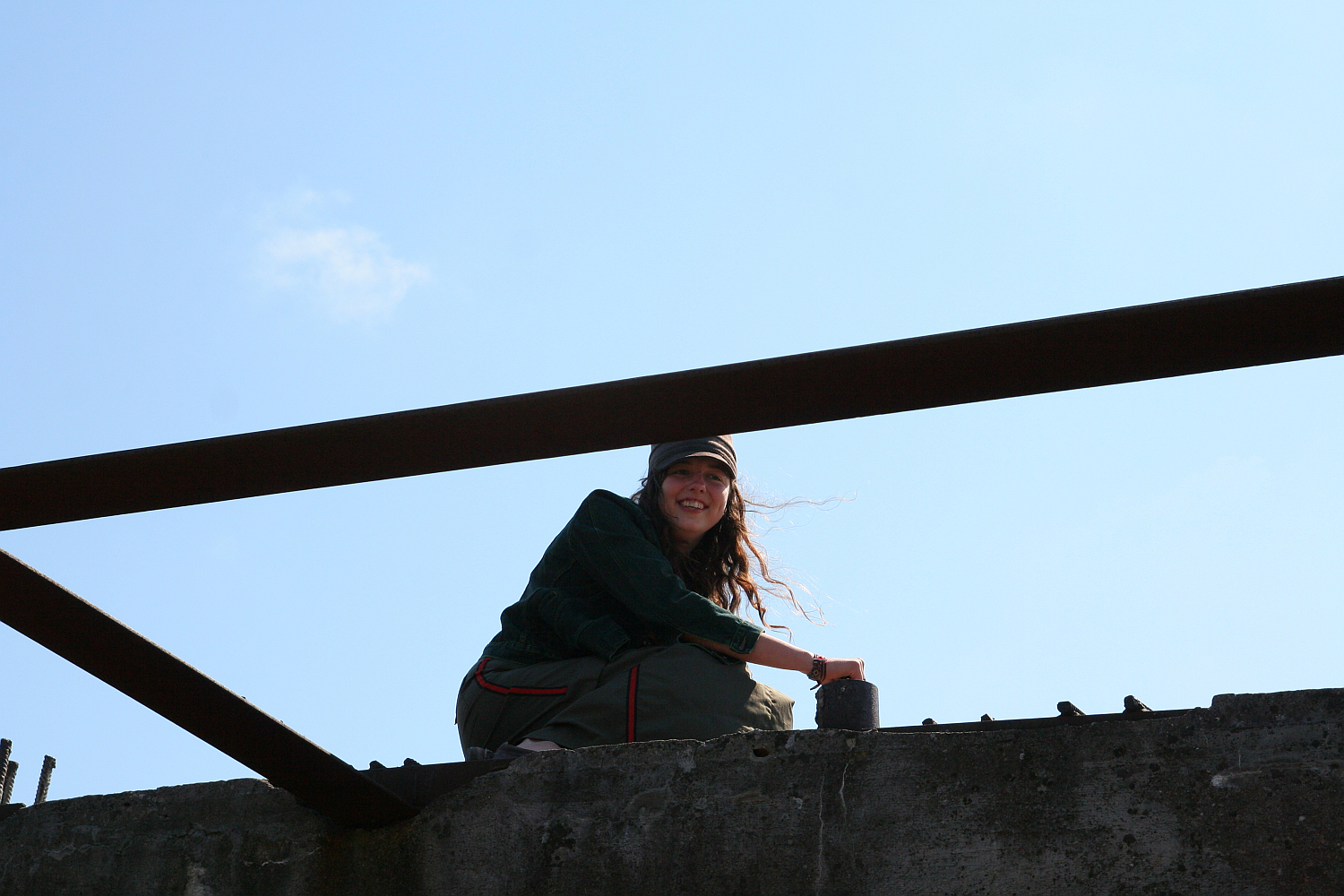 A smiling person sits atop a concrete structure under a clear blue sky, resting her arms on a rusty metal beam. Her casual attire and relaxed posture suggest she's enjoying a moment of leisure in an industrial