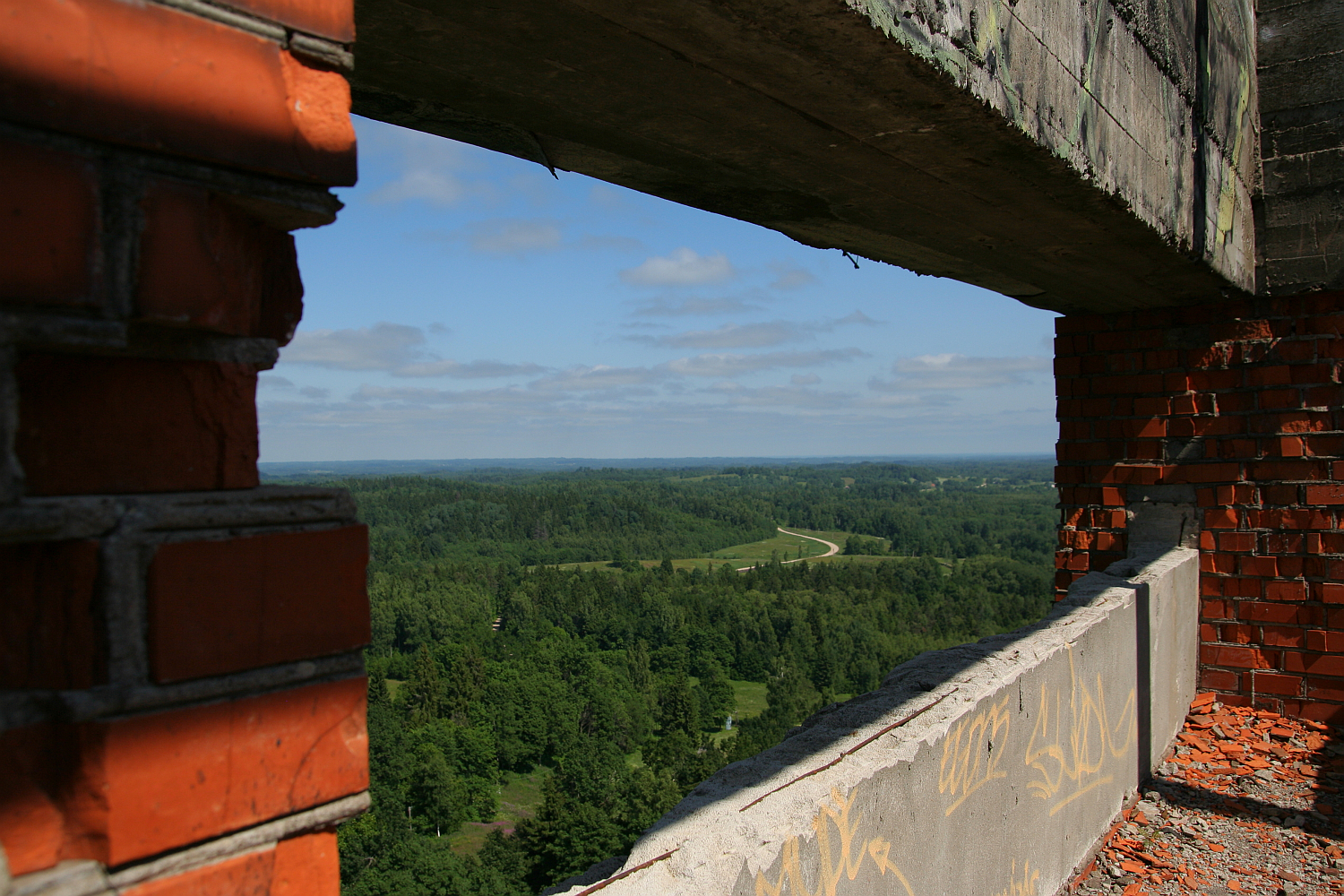 A view from a dilapidated structure with exposed brickwork overlooking the lush green forest landscape of Gaiziņš and winding road, under a clear blue sky.