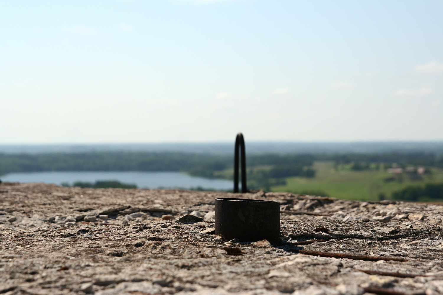 A metal loop protrudes from weathered concrete with a scenic view of Gaiziņš, a peaceful lake, and green landscape blurred in the background, under a clear blue sky.