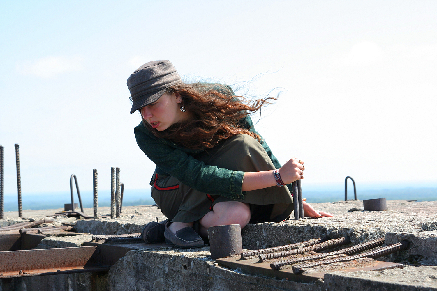 A woman with long, flowing hair and wearing a gray hat and green jacket crouches on a concrete structure near Gaiziņš. She is holding a metal rod and surrounded by rebar and metal components. The sky is clear with a faint horizon in the background. She appears focused on her task.