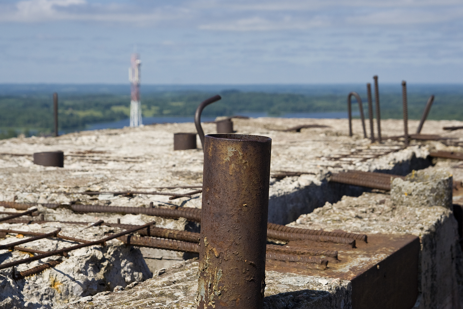A close-up of a rusted metal rod protruding from the crumbling concrete of an abandoned, unfinished building. The foreground is filled with more exposed rebar and deteriorating concrete. In the distance, there's a blurred view of a green forested landscape and Gaiziņš standing tall against the horizon.