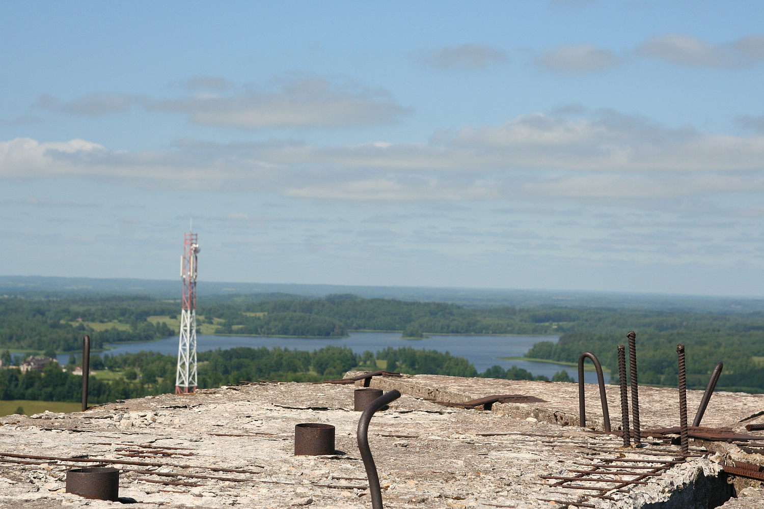 A wide-view landscape image showing a concrete surface with exposed rebar in the foreground, overlooking a lush green expanse with scattered trees and a lake in the background. A tall communication tower stands to the left amidst the greenery near Gaiziņš. The sky is blue with some clouds.