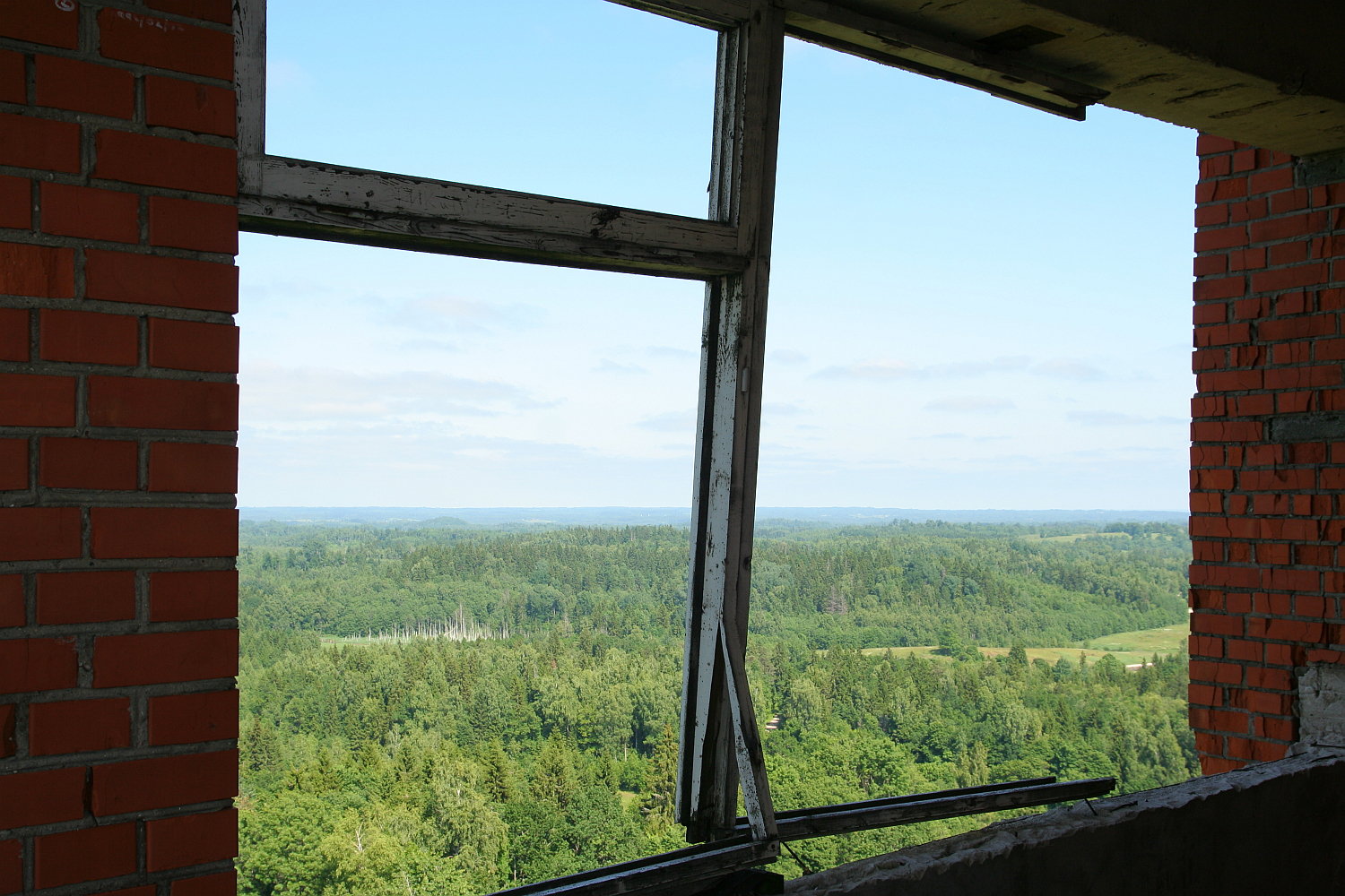 A view through a broken window frame in a red brick structure, overlooking the lush green landscape of Gaiziņš under a clear blue sky.