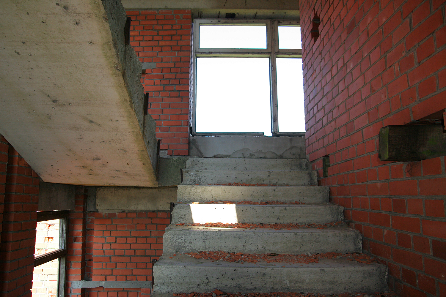 A gritty, unfinished staircase with bare red brick walls and exposed concrete surfaces leading to a brightly-lit window atop Gaiziņš, exuding a raw, industrial atmosphere.