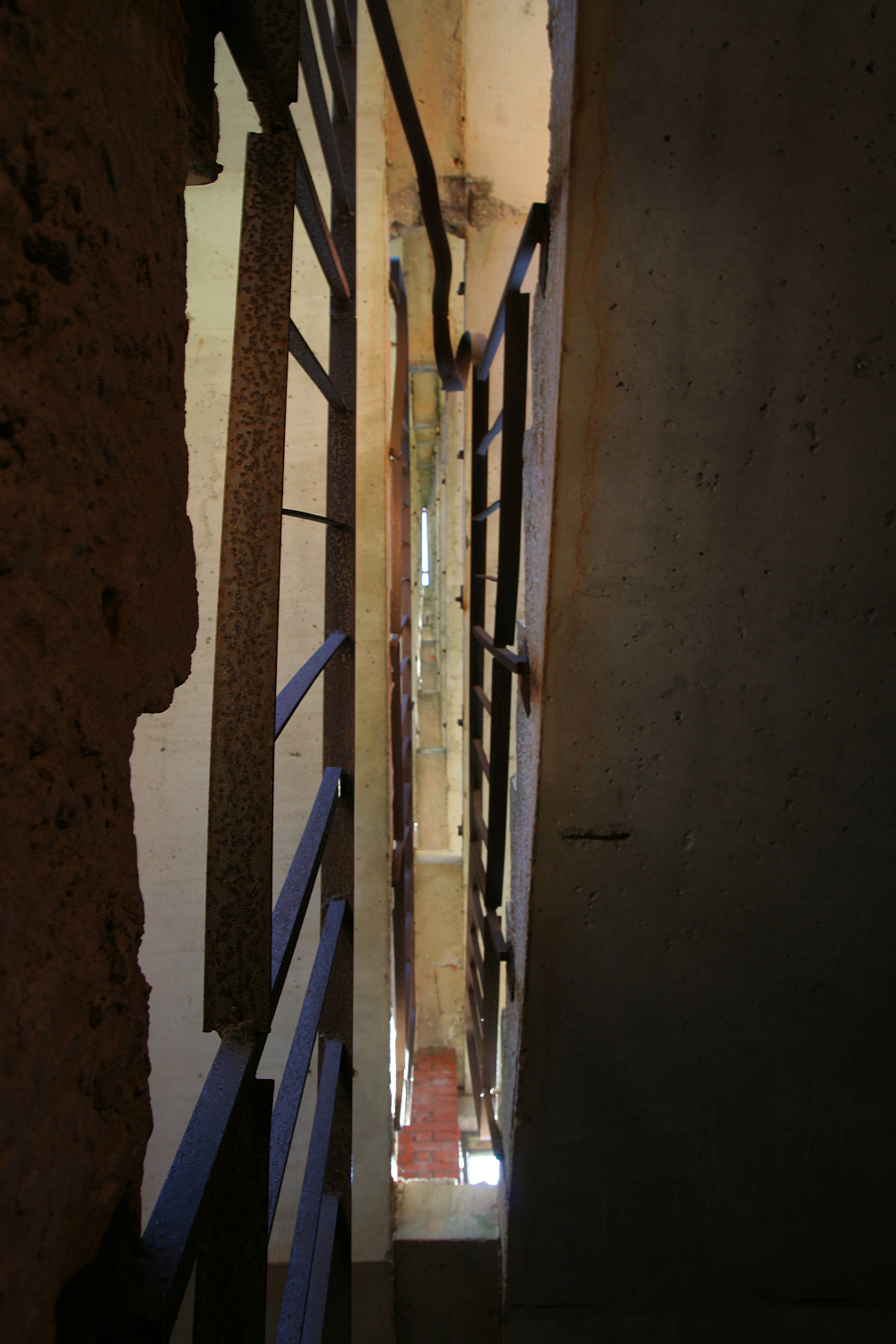 A dimly lit stairwell with rusted metal banisters ascends between weathered concrete walls, while narrow slits of light peek through, hinting at an urban, gritty Gaiziņ