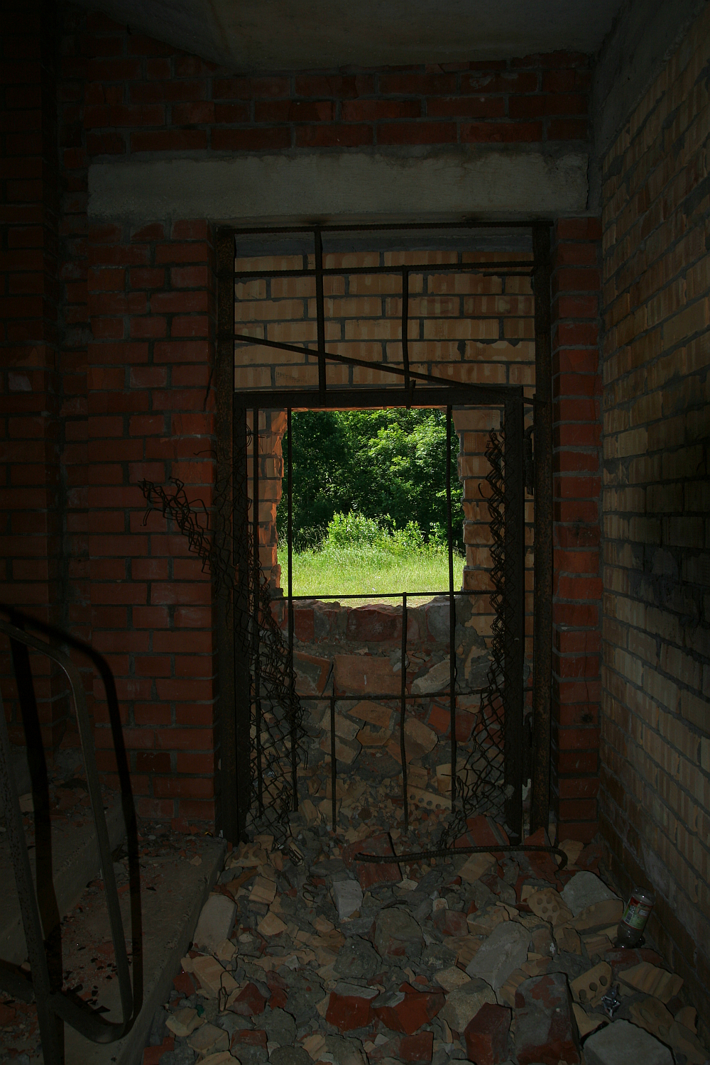 A dilapidated room with crumbling brick walls and scattered debris leads to a rusted, broken metal gate, framing a view of Gaiziņš's lush greenery outside, highlighting contrasts of