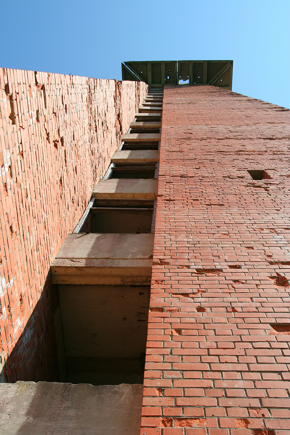 A towering view from the base of an imposing brick structure at Gaiziņš, showcasing a series of protruding concrete steps ascending to a platform under a clear blue sky.