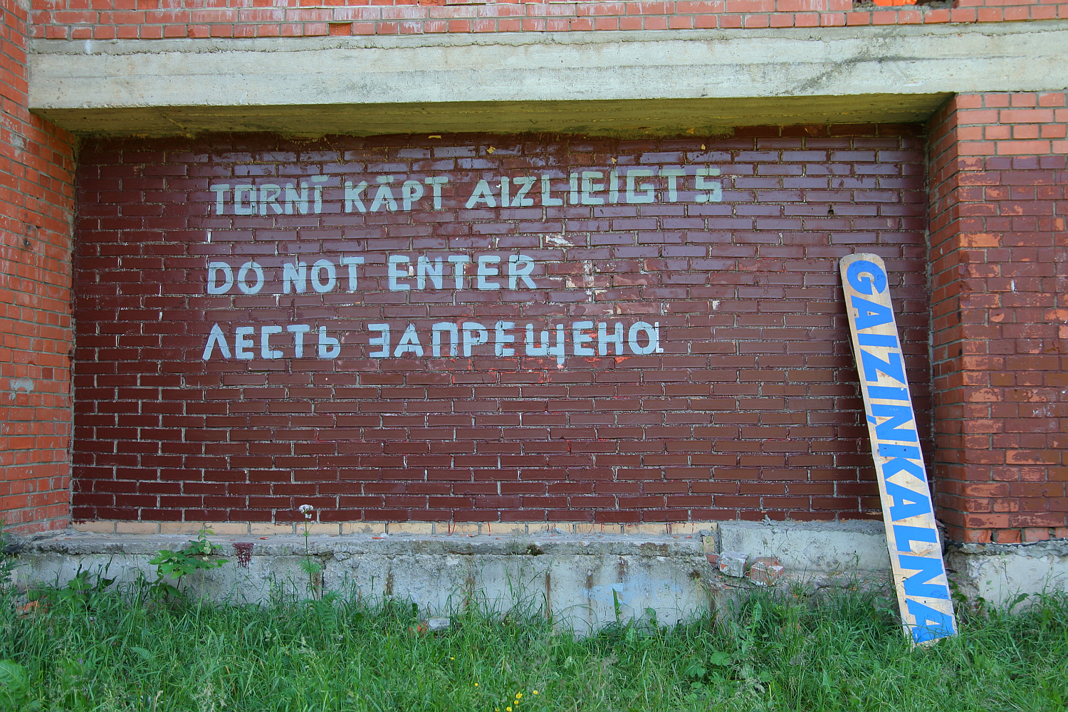 A brick wall with a sign in multiple languages, including Latvian, English, and Russian, reads, "TURNI KĀPT AIZLIEGTS," "DO NOT ENTER," and "ЛЕСТЬ ЗАПРЕЩЕНО," indicating that entry is forbidden. A blue and white sign leaning against the wall features the word “GAIZINKALNS.” Grass is
