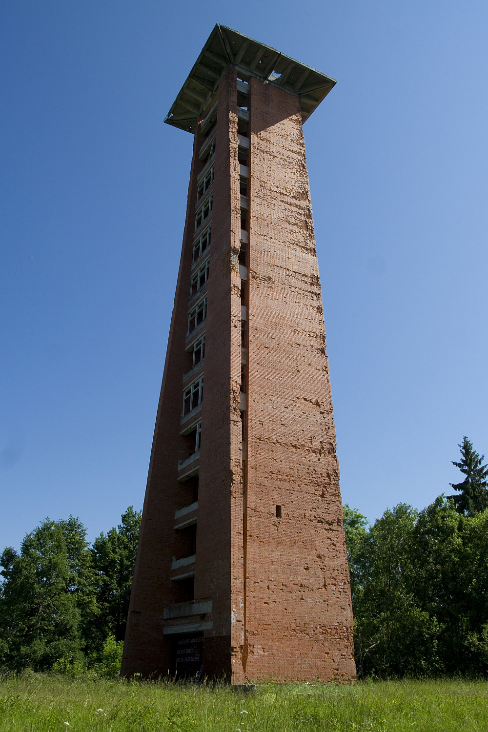 A tall, narrow, red brick tower with a pyramidal top and a slightly overhanging green roof stands solitary against a clear blue sky, surrounded by lush Gaiziņš greenery
