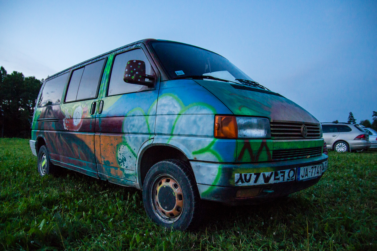 A colorful van, adorned with vibrant graffiti and abstract designs, is parked on a grassy field near a forested area at dusk. The scene harkens back to the 2013 Give and Get festival. The sky transitions to night, casting a blue hue, while a silver car is partially visible in the background on the right side.
