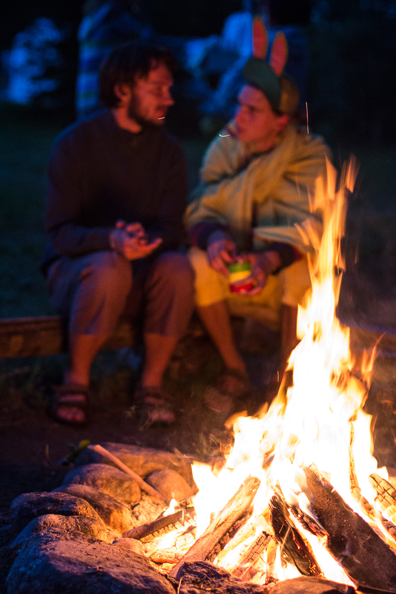 Two people sit by a campfire at night, engaged in conversation. The person on the right wears a yellow onesie with rabbit ears, while the person on the left wears casual clothes. The warm firelight illuminates them, creating a cozy atmosphere against the dark background of the 2013 Give and Get festival.