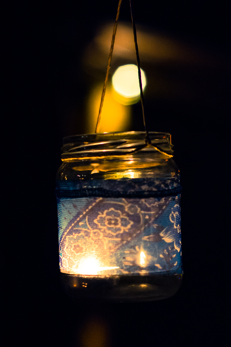 A glass jar with a lit candle inside hangs by a thin string against a dark background. Adorned with decorative blue lace fabric, the jar casts intricate floral patterns when illuminated by the candlelight, creating a warm, soft glow. A blurred bright spot appears in the background of this 2013 Give and Get festival scene.