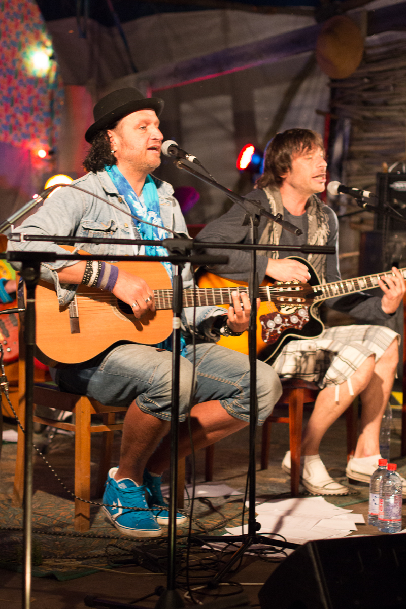 Two male musicians perform on stage with acoustic guitars during the 2013 Give and Get festival. The man on the left wears a black hat, blue scarf, denim jacket, and jeans with blue sneakers. The man on the right sports a grey shirt and plaid shorts with black boots. Both sing into microphones as they play their guitars.