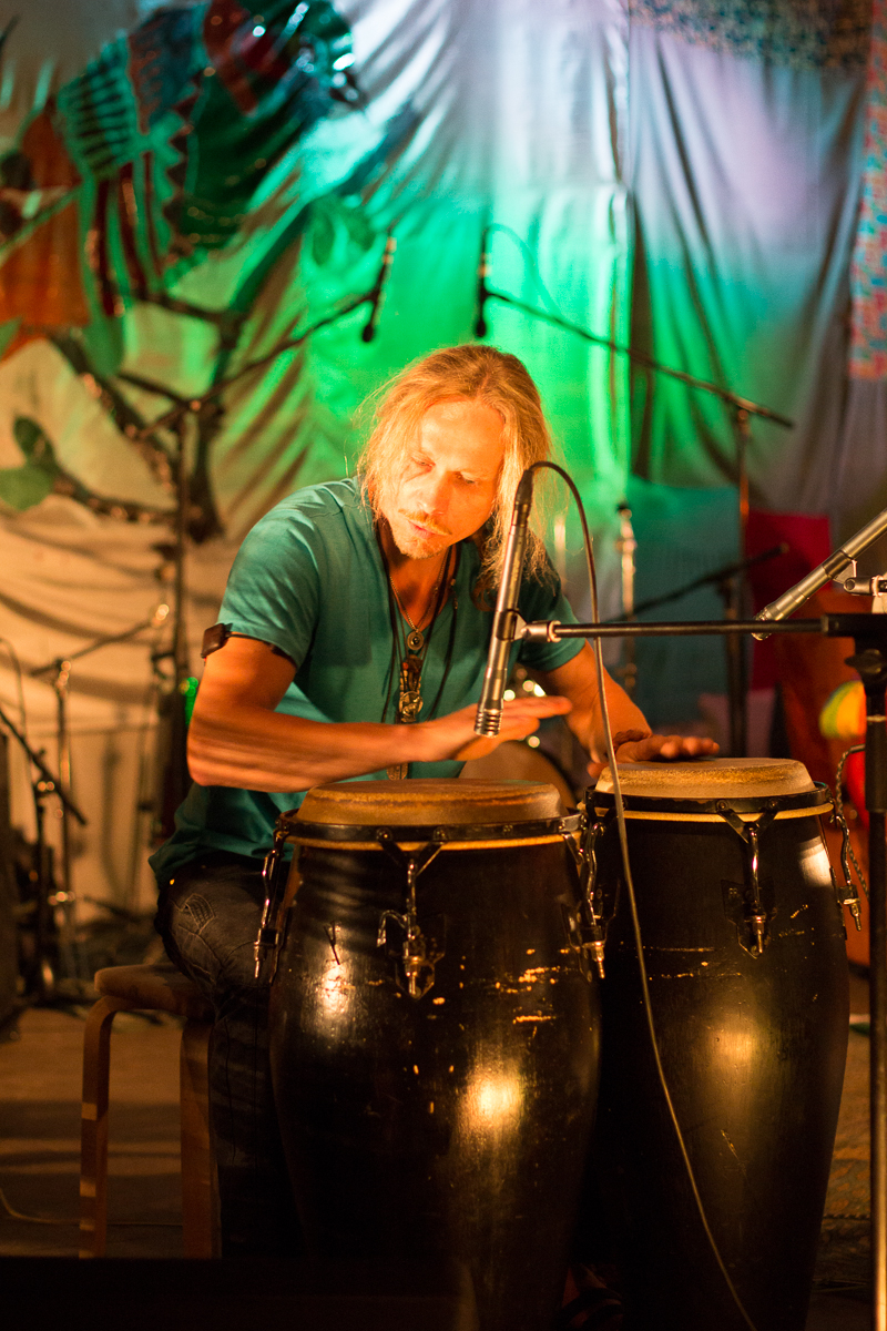 A man with long blonde hair and a blue shirt plays two large conga drums in a colorful, dimly lit setting at the Give and Get 2013 festival. A microphone is positioned near the drums, capturing the sound. The background features vibrant, multi-colored decorations and stage lighting, with additional drums and musical equipment visible.