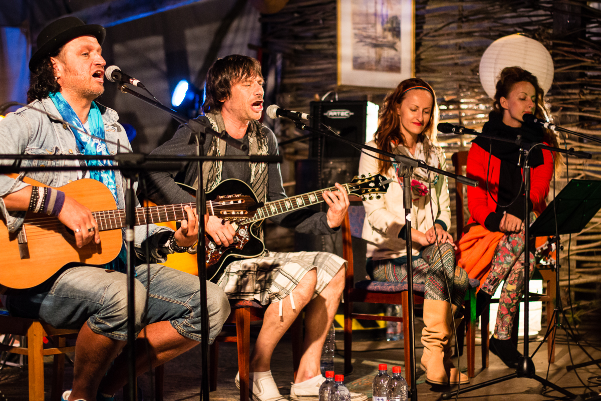 Four musicians are performing on stage at the 2013 Give and Get Festival. Two men on the left are playing acoustic guitars. One wears a hat, scarf, and jean shorts; the other sings passionately into a microphone. Two women on the right sing into microphones; one in a white jacket and patterned leggings, and the other in a red top and black scarf.