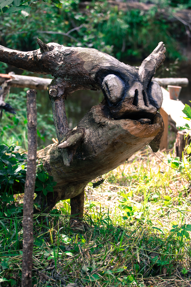 A weathered wooden sculpture resembling a fantastical creature stands on grass. It has large, round eye-like protrusions and horn-like extensions on its head. The setting, reminiscent of the 2013 Give and Get festival, is a lush green area with blurred foliage and a small body of water in the background.