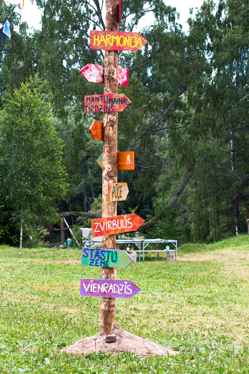 A colorful wooden signpost stands in a grassy clearing, surrounded by trees. The signpost, from the 2013 Give and Get festival, is adorned with multiple arrow-shaped signs in vibrant colors, with text in Latvian pointing in different directions. A picnic table and a few objects are visible in the background.