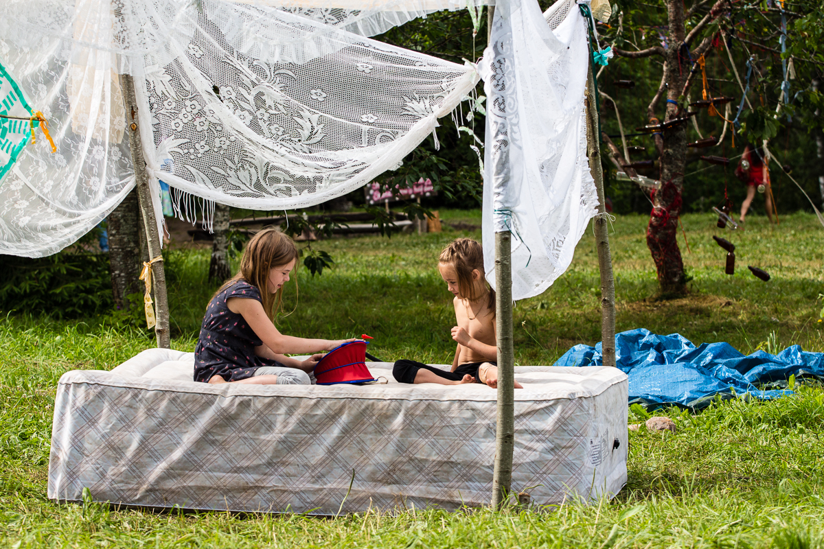 Two young girls sit on an outdoor mattress under a makeshift canopy made of lace curtains and wooden poles. One girl holds a red toy sailboat, while the other watches. The scene is set in a grassy area with scattered items and greenery in the background, reminiscent of a 2013 festival vibe.