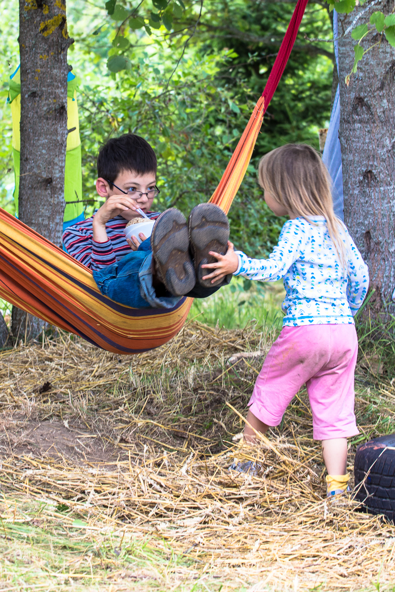 A boy with glasses is lying in a colorful hammock holding a cup, wearing a striped shirt, jeans, and brown shoes. A young girl in a white star-patterned shirt and pink pants stands next to him with her arm extended under a tree in a grassy area. Her clothes and feet are dirty from the 2013 Give and Get festival.