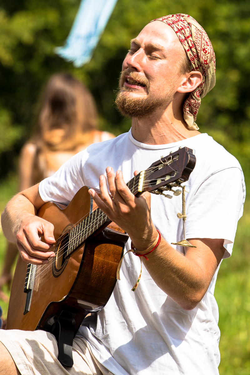 A man with a beard, wearing a white shirt and a patterned bandana, plays an acoustic guitar outdoors at the Give and Get Festival 2013. He appears to be passionately singing with his eyes closed. The background features greenery and an out-of-focus person, and a blue vertical flag or banner is partially visible.