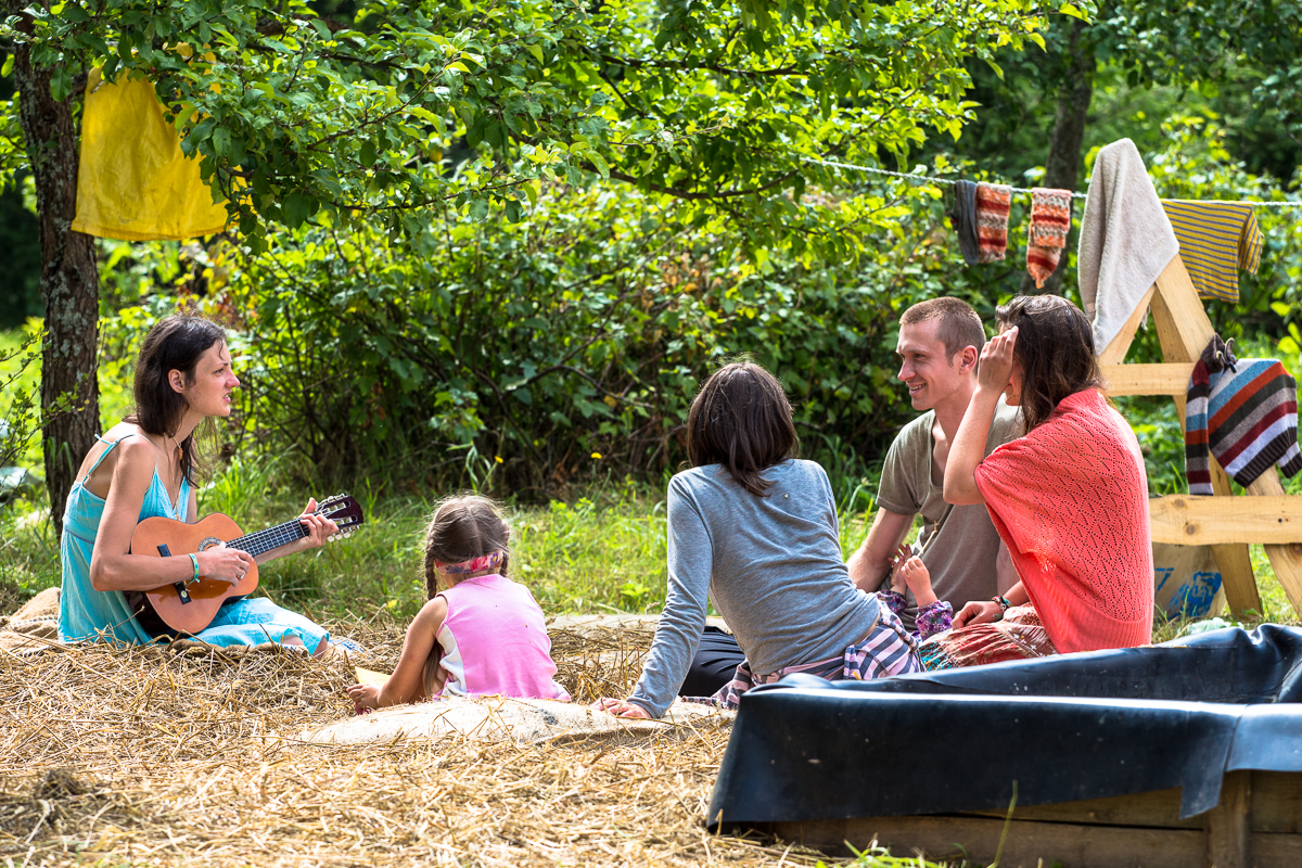 A group of five people, including two children and three adults, relaxes outdoors on a grassy area with hay at the Give and Get festival 2013. One adult plays a small guitar while the others listen or chat. Towels hang on a line behind them, and trees provide shade. A large plastic container is visible in the foreground.