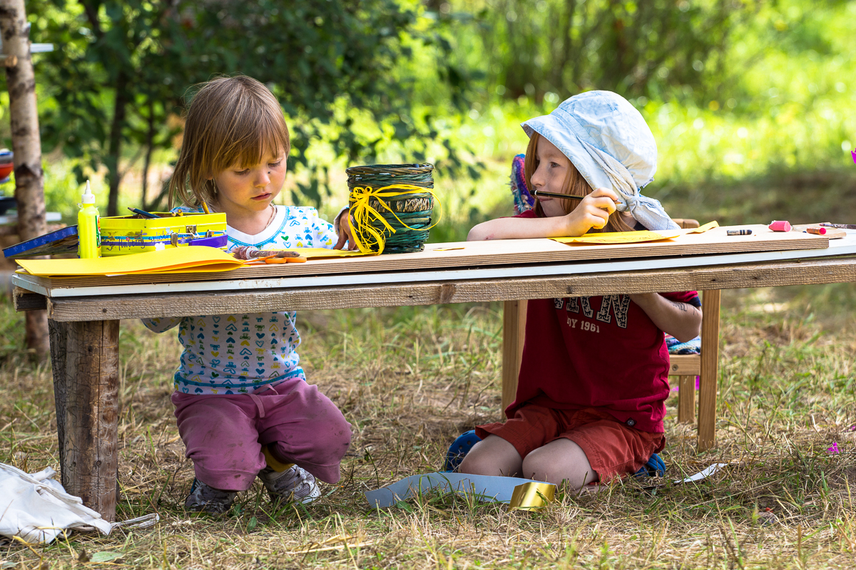 At the 2013 Give and Get festival, two young children are seated outdoors at a low wooden table engaging in art activities. The child on the left wears a white shirt with blue patterns and pink pants, while the child on the right sports a blue hat, red shirt, and shorts. The table is covered with art supplies, including papers and markers.