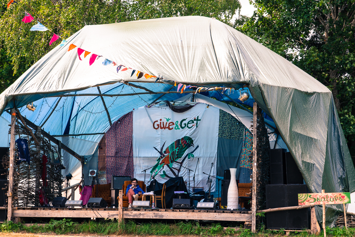 A covered outdoor stage with a "Give & Get" backdrop, reminiscent of the 2013 Give and Get festival. The stage is set with musical instruments and equipment, including a drum set and speakers. A person in a blue shirt sits before the backdrop. Colorful triangular flags hang above, framed by trees in the background.