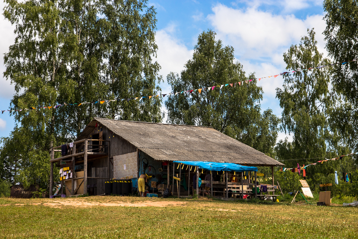 At the Give and Get 2013 festival, an outdoor market stall is set up under a rustic wooden canopy with a corrugated metal roof. Colorful bunting hangs above, and items are displayed along the wooden structure. Trees and a partly cloudy blue sky create a festive, rural atmosphere.