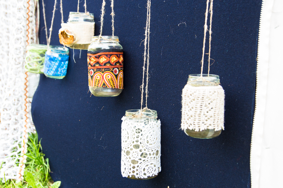 A display shows six glass jars hanging by twine against a dark blue background. Each jar is decorated with a different patterned fabric or lace. In the bottom left corner, a small patch of greenery is visible, along with a crochet piece draped partially in view, reminiscent of the 2013 Give and Get festival.