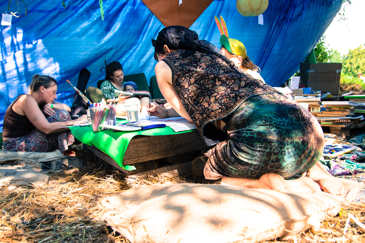 A group of people gathered under a blue tarp canopy at the 2013 Give and Get Festival, engaged in various activities. One person kneels at a low table covered with art supplies, while others sit on mats around the table. Sunlight filters through the trees, casting dappled shadows on the scene.