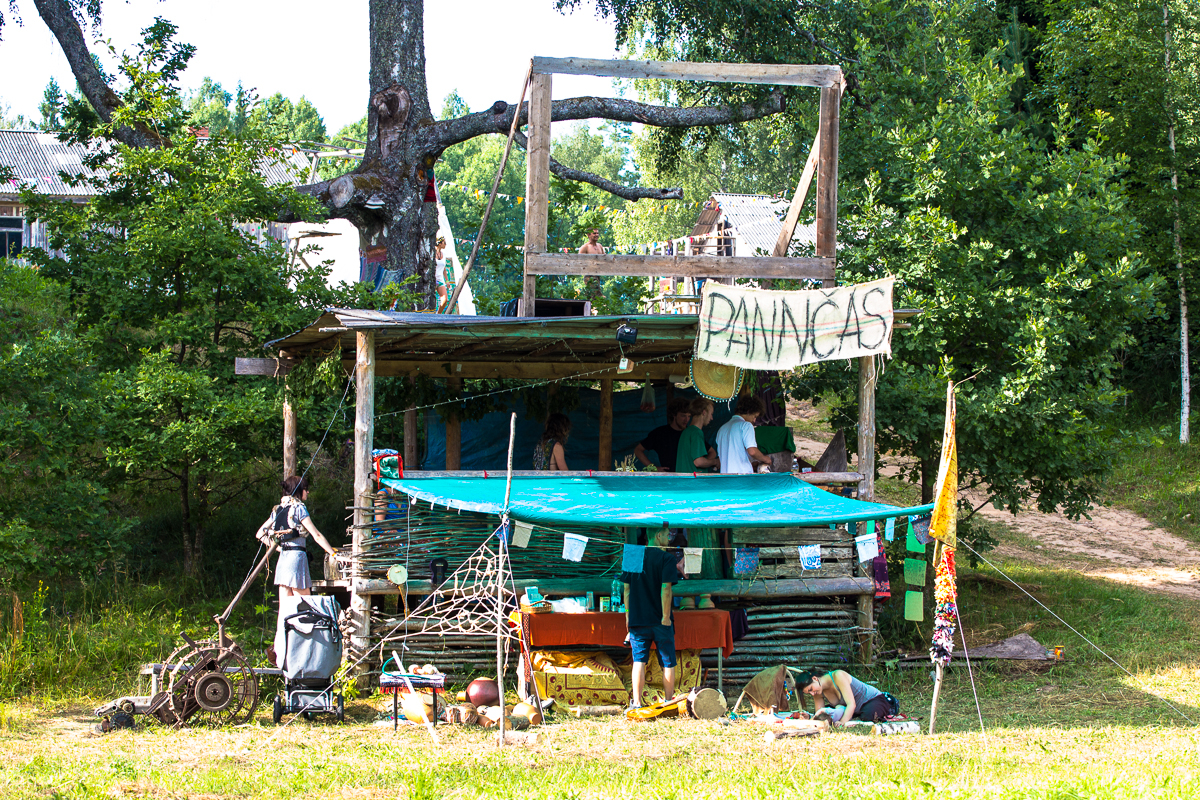 A wooden shack surrounded by trees with an upper deck, a light blue tarp for shade, and a sign reading "PANNAS" in large letters. People are gathered around and inside the shack, engaging in various activities at the 2013 Give and Get festival. The scene is bright and sunny, with grassy ground below.