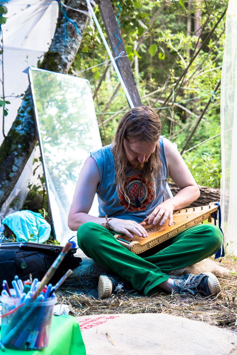 A person with long hair and a beard plays a stringed instrument while sitting on a blanket outdoors. They wear a sleeveless shirt with a circular design and green pants. Surrounding them are art supplies, a large mirror, and trees in the background. The scene evokes the relaxed, artistic vibe of the 2013 Give and Get festival.