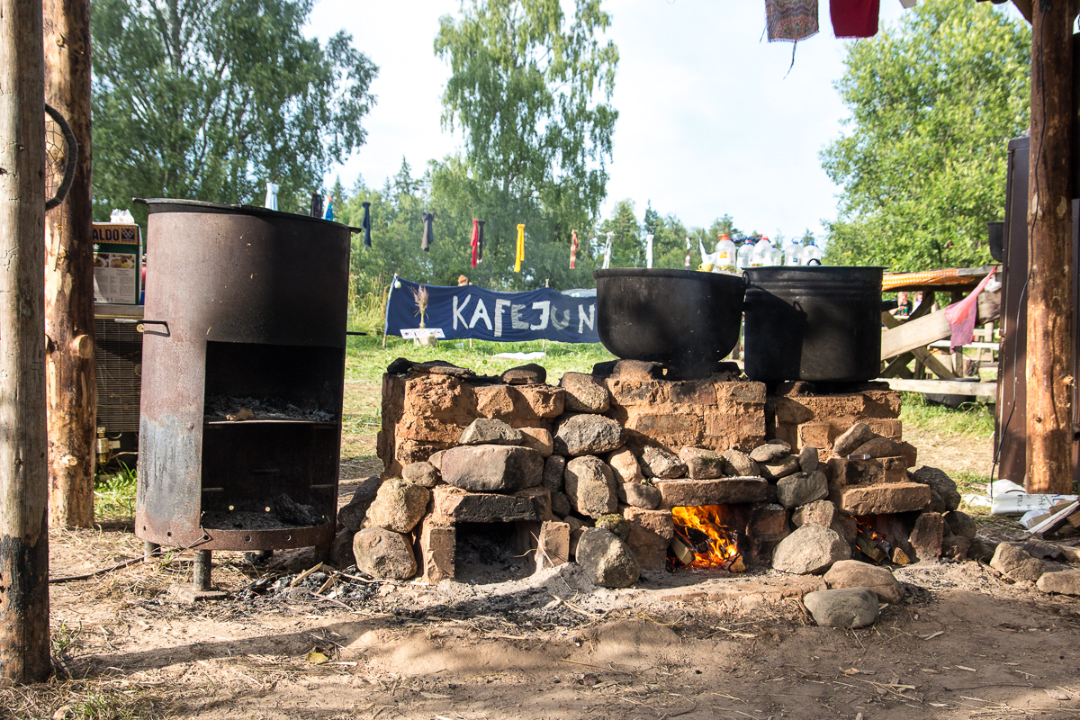 An outdoor makeshift kitchen at the 2013 Give and Get festival features a large metal stove and two big pots on a stone and brick hearth. A small fire burns underneath, with wooden shelter frames and drying laundry visible. In the background, a banner reading "KAFE3" is surrounded by trees and grassy landscape.