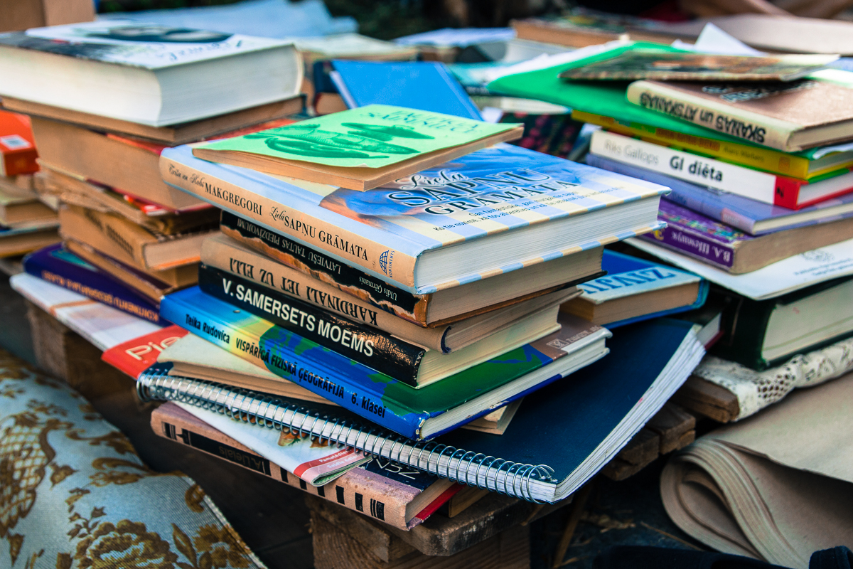 A large pile of books stacked haphazardly on a wooden surface. The books have various colorful covers and titles in different languages, including Latvian and English. Some spiral notebooks are also interspersed amongst the books from the 2013 Give and Get festival. The background is slightly blurred, showing more books and fabric.