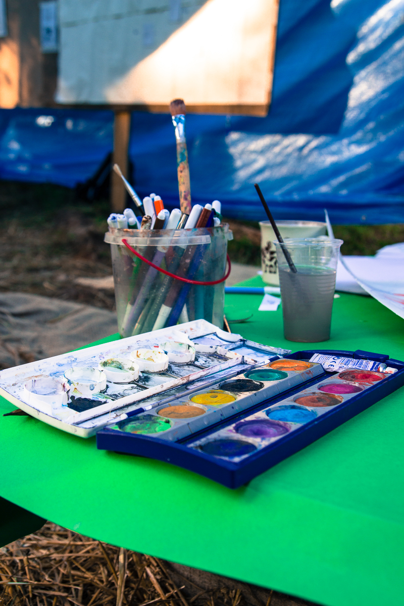 A table outdoors is covered with art supplies, reminiscent of the creative spirit at the Give and Get festival 2013. In the foreground, watercolor palettes burst with various colors. Nearby, a plastic cup holds water and paintbrushes, while a second cup contains murky water with a brush in it. Background includes a blue tarp and a whiteboard.