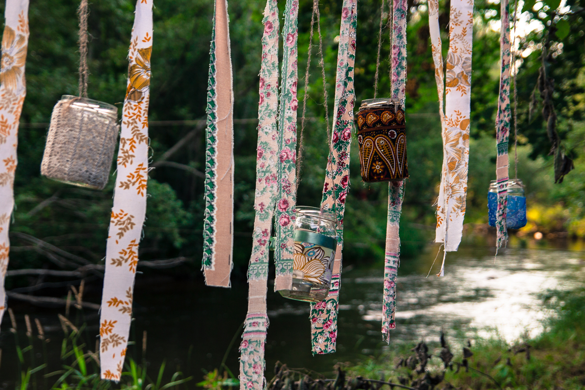 Glass jars and fabric strips hang from tree branches near a peaceful river, creating a whimsical and artistic atmosphere reminiscent of the 2013 festival. The jars are painted with colorful designs, some filled with small objects, while the fabric strips display various patterns and colors.