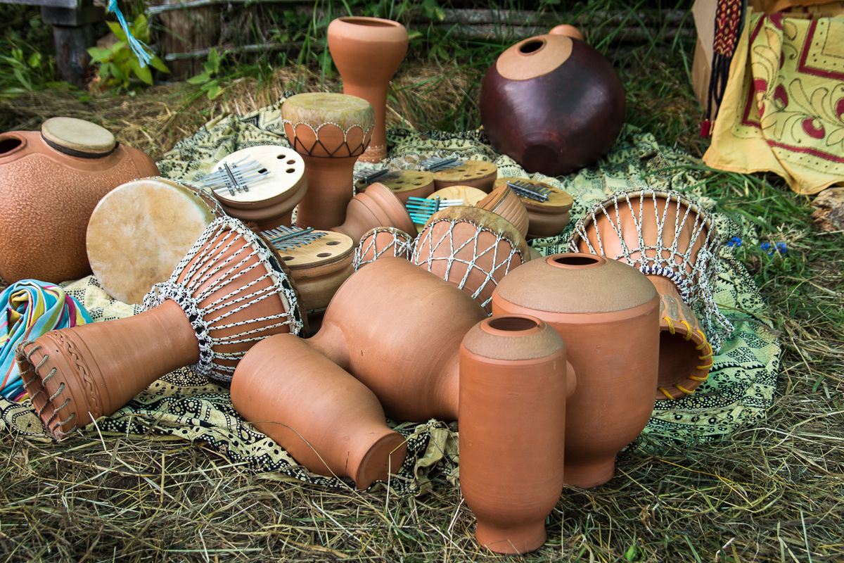 Various clay and wooden musical instruments, including drums, kalimbas, and jars, are laid out on a patterned cloth on grass. The instruments feature intricate designs and are used in traditional music. The setting appears rustic, with wooden planks and greenery visible in the background of the 2013 Give and Get festival.
