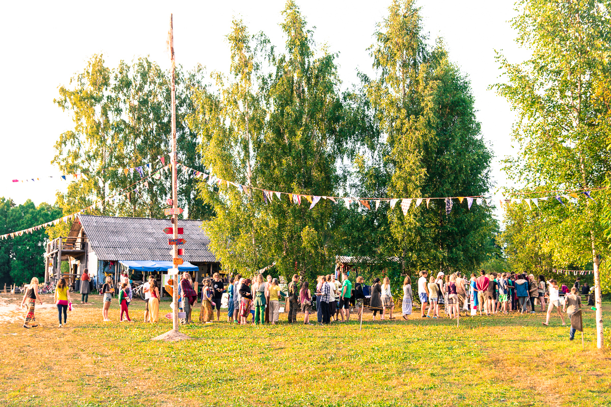 A lively outdoor scene at the 2013 Give and Get festival in a rural area, featuring a crowd of people gathered under colorful bunting flags strung between tall trees. A rustic wooden building is visible in the background. The event takes place on a sunny day, creating a warm and festive atmosphere.