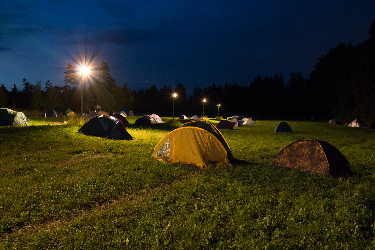 A nighttime scene of a campsite during the 2013 Give and Get festival, with multiple tents pitched on a grassy field. Several tall lampposts illuminate the area with a warm glow, casting light and shadows on the tents and grass. The sky is dark blue, indicating it is well into the night, with trees silhouetted in the background.