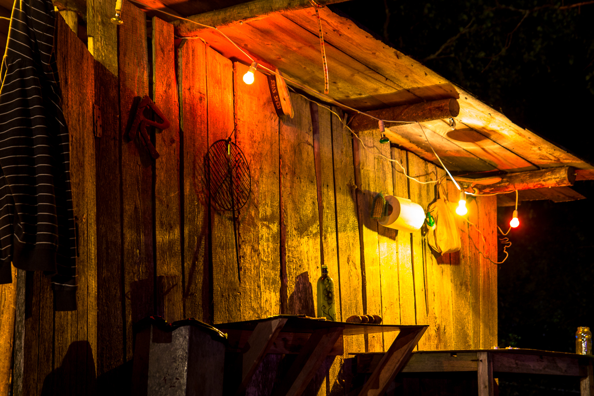 A rustic wooden shack is dimly lit by colorful string lights, including red, yellow, and green bulbs. A striped shirt from the 2013 Give and Get Festival hangs on the left wall, and various items, including a wire rack and a plastic bottle, are attached to the walls. The scene is set against a dark, night-time background.