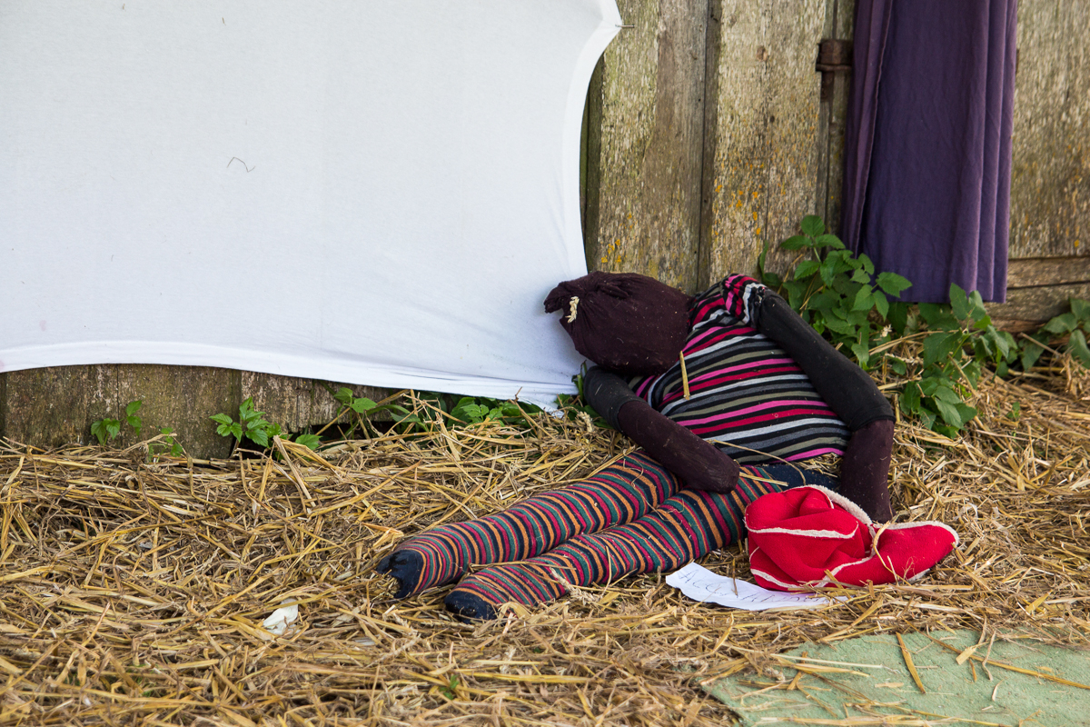 A homemade scarecrow, made from a fabric doll dressed in black, red, and striped clothing, leans against a weathered wooden wall. Surrounded by hay and green plants nearby, it evokes memories of the 2013 Give and Get festival. A white sheet is positioned in the background with a pair of red shoes near its feet.