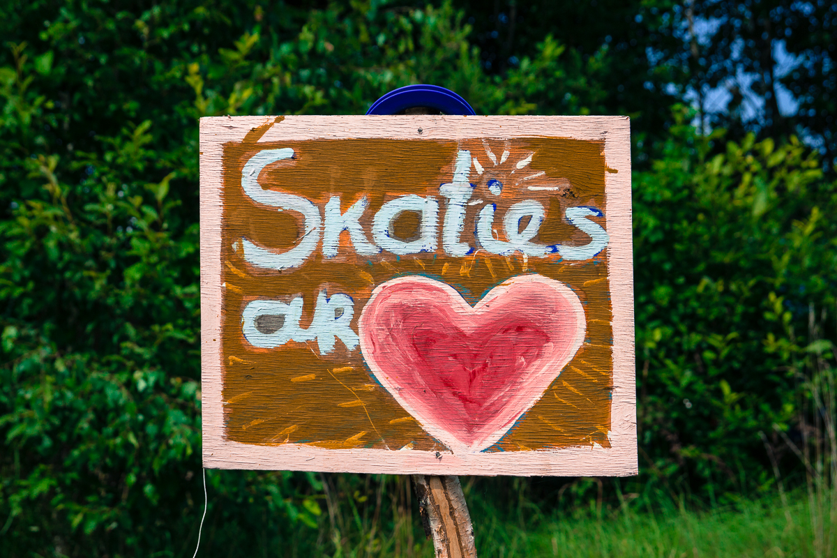 A handmade wooden sign is displayed against a backdrop of lush greenery at the 2013 Give and Get festival. The sign reads, "Skaties ar" in white, with a large red heart painted below the text. It has a rustic appearance, with visible brushstrokes and a brown background.