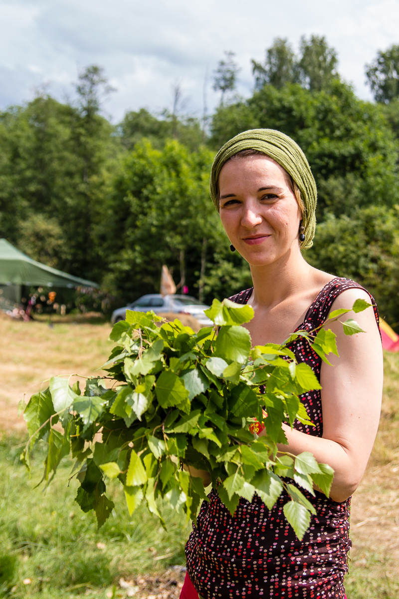 A woman with a calm expression stands outdoors holding a bunch of fresh green leaves. She wears a sleeveless black and red patterned top and has a green headscarf. Behind her, there are trees, a vehicle, and a green canopy tent, evoking the vibrant spirit of the Give and Get Festival 2013 in this forested, camp-like setting.