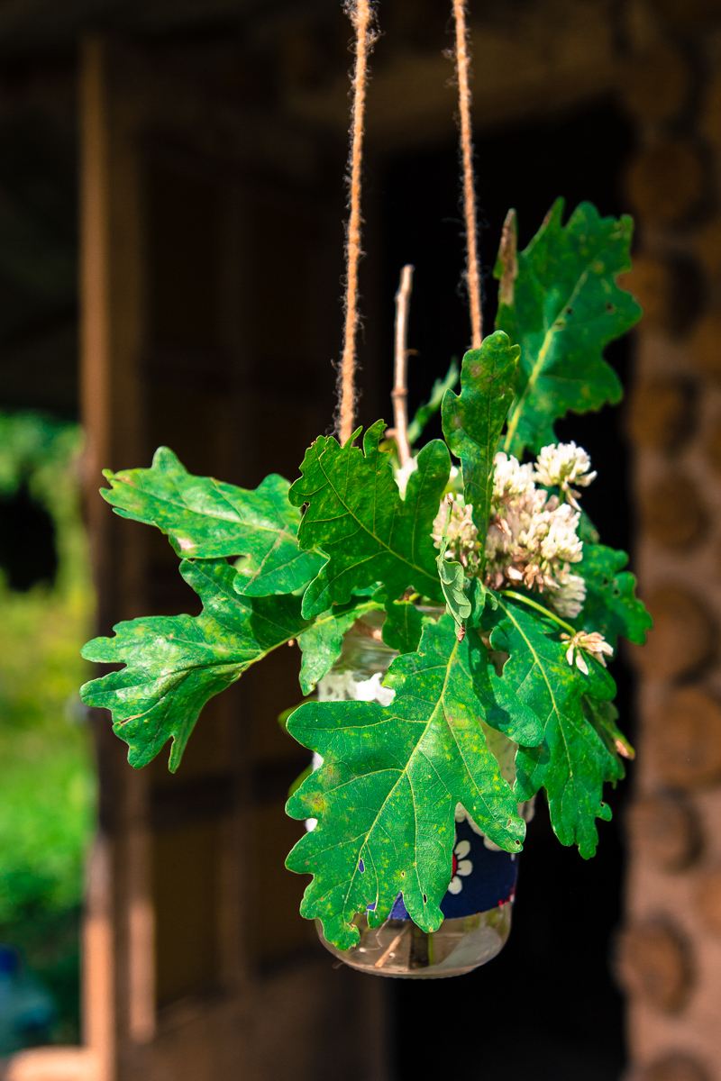 A small glass jar wrapped with twine hangs from a rope, containing a bouquet of green oak leaves and light pink flowers. The background is slightly blurred, showing a rustic wooden structure with a doorway, allowing sunlight to filter through and illuminate the hanging jar at the 2013 Give and Get festival.
