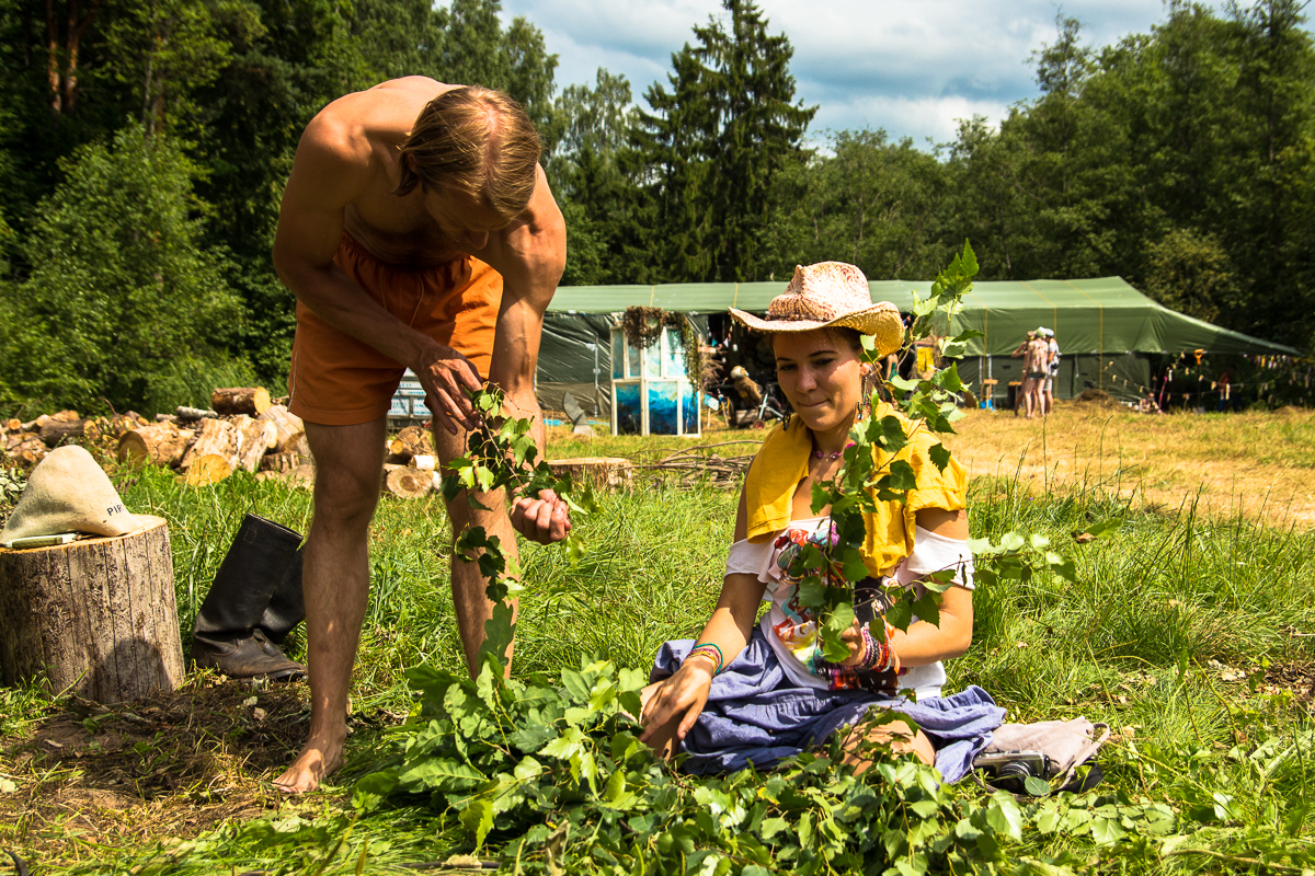 A man in orange shorts and a woman in a yellow top and blue skirt with a cowboy hat are crafting greenery wreaths outdoors. They are surrounded by lush trees and greenery at the Give and Get festival, with people standing near a large green tent in the background under a partly cloudy sky.
