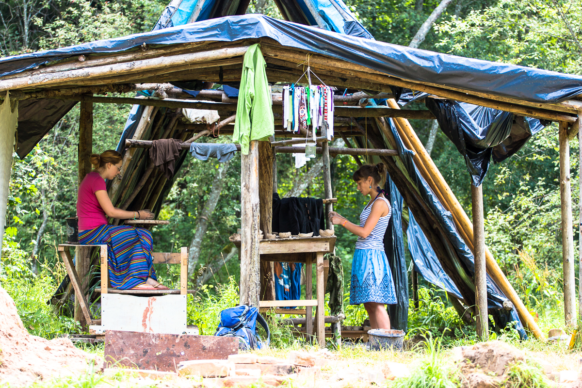 At the 2013 Give and Get festival, two women work under a makeshift wooden shelter with a tarp roof in a green outdoor setting. One woman sits weaving while the other stands, painting or staining cloth. Various clothes hang to dry above them, with trees and foliage visible in the background.