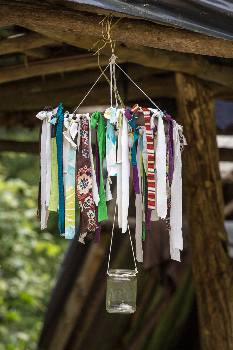 A homemade hanging mobile, reminiscent of the 2013 Give and Get festival, features numerous colorful fabric strips tied to a circular frame suspended by strings. A clear glass jar hangs below the frame, adding charm. The background reveals a rustic wooden structure with a green outdoors visible beyond.