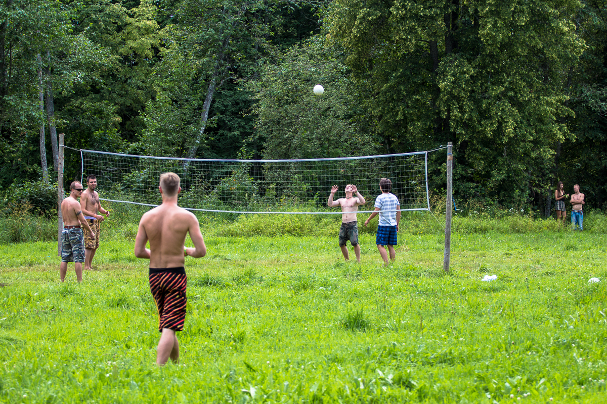 During the 2013 Give and Get festival, a group of shirtless people are playing volleyball on a grassy field surrounded by trees. The scene shows a net and several players actively engaged in the game, with two specifically focused on hitting the ball while others are positioned around them. Two individuals watch from the background.