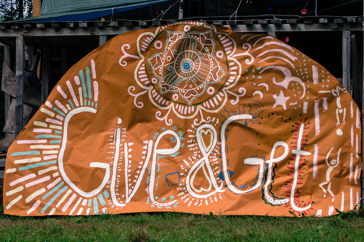 A large, orange banner with intricate, white and blue designs featuring swirls, stars, and an eye symbol at the top. The banner bears the painted words "Give&Get" in large, stylized white and red lettering. Displayed outdoors on a rustic wooden structure above green grass, it heralds the Give and Get festival 2013.
