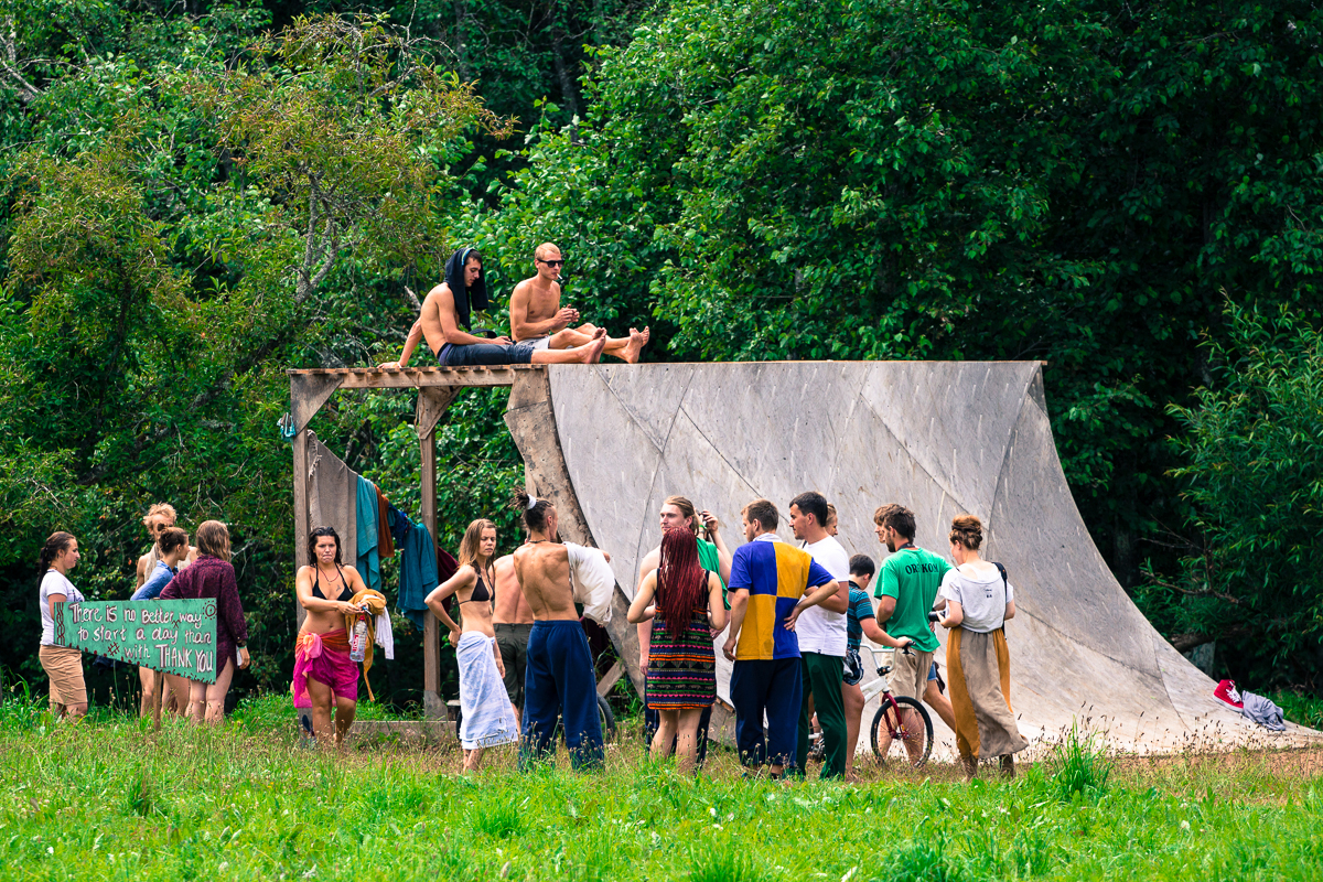 A group of people gather outdoors near a wooden ramp structure at the Give and Get festival in 2013. Some stand and chat, while others sit on top of the ramp. One individual holds a colorful sign with text on it. The scene is set in a lush, green environment with trees in the background and grass underfoot.